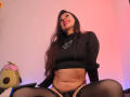 AmaraMirage with long dark hair, black crop top, and black lace panties, sitting with legs apart, showing her pussy, wearing black stockings, smiling with red lipstick.