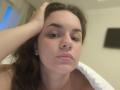 Dreia with brown hair, slightly parted lips, and hand in hair, looking at camera, showing soft pink nipples under white pillow. Blurred background. Casual, slightly blurry photograph.