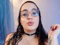 StacyMillerr with wet black hair, glasses, and a black choker, pouting with pink lips, has a tattoo on her right hand, showing off her nipples.