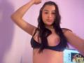 IrisBlair with long dark hair, arm raised, wearing a black bra that exposes her right tit. She has light skin and a neutral expression. Computer screen in the background.
