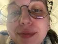 Sanella's face, showing her nose and lips with a slight smile, wearing round glasses with reflective lenses, and a blue and white fuzzy sweater, in a close-up shot.