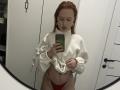 JannaCorsaro with red hair, white turtleneck sweater, red thong, lifting sweater, exposing her tits and belly tattoo, taking mirror selfie, holding green phone, white room.