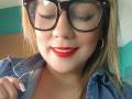 CamilaNoer has red lipstick, black glasses, and a denim shirt, with blonde hair and hoop earrings. Her tits are not visible. She's smiling with red lipstick.