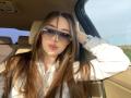 JaneKrover with long brown hair, wearing oversized sunglasses and a white shirt, has her hand in her hair, showing her pink lipstick and silver watch, sitting in a car.