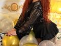 Niferttiti with curly red hair, wearing a black fishnet top and black ruffled skirt, kneeling on a bed with gold and clear balloons, showing her tattooed thigh.