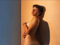 Nude GagingMolly with short curly brown hair, small tits, and a cute ass, standing against a wall, arms crossed, looking over shoulder, sunlight casting shadows.