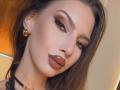 LucyGrace with glossy brown lips, thick winged eyeliner, brown hair, gold earrings, and a black choker, looking seductively at the camera with a slight smile.