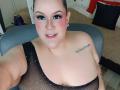 GiiannaMorrys with pink blush, black mesh top, visible large boobs, tattoo on right shoulder, nose ring, smiling, brown hair, blue eye makeup, taking selfie, gray chair.