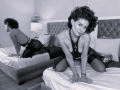 Schwarz-weißes Foto von LilaaSalmon auf einem Bett, die ein durchsichtiges Kleid mit Perlen trägt, wobei ihre Brüste sichtbar sind, sie kniet und ihren Hintern hebt, die Spiegelung zeigt ihren Rücken..
