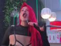 AlissaBrown with bright red hair, black sheer top, exposed tits, tongue out, holding a dildo to her mouth, in a modern room with white, red, and green lights.