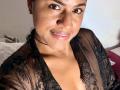 IvannaDuran69 with tan skin, dark eyebrows, and shiny pink lips, wearing a black lace robe, showing her small tits, smiling at the camera. Her face is centered, top view.