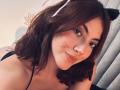 SpencerLe with cat ears, brown wavy hair, black strappy top, slight smile, fake whiskers on cheek, cute, light skin, medium-sized tits, black furry cat ear headband, casual selfie.