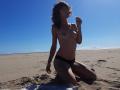 Topless Aiyana with small tits, black panties, kneeling on sandy beach, holding lollipop, blue sky, sunlit, wind in brown wavy hair, tan skin, beach towel beneath.