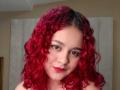 RosalieVittoria has red curly hair, brown eyes, and red lipstick, looking at the camera with a slight smile, showing her cute face and soft skin.