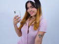 AbbyJanes with blonde to brown ombre hair, pink shirt, white headband, holding phone, smiling, pink eyeshadow, small tits, tattoo on right arm, pink nails, holding phone.