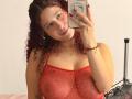 ValeryScot with curly red hair, wearing a red mesh top, showing her large tits, holding a phone, smiling, red nails, gold necklace, and bracelet, in a selfie.