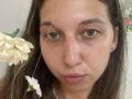 Sanella's face with brown eyes, slightly shiny skin, and brown hair, wearing a small earring, holding white flowers in a selfie, with a visible light reflection on her forehead.