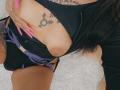 DenyChantal's tits are exposed, showing a brown areola, with pink nails, black top, purple straps, and tattoos, including a gender symbol on her chest.