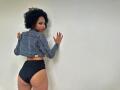 ChantalHaneke with curly black hair, wearing a denim jacket and black high-waisted underwear, looking over her shoulder, hands on a white wall, showing her ass.