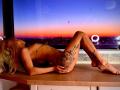 NayaBliss, nude, with tattoos on thigh and side, laying on wooden table, back arched, legs bent, blonde hair, sunset through window, city lights in background, night sky.