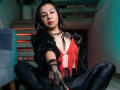 MissCharlotteJolie with black lace gloves, red fringe top, black leather pants, and black feather jacket, sitting cross-legged, wearing a chain necklace, looking at the camera.