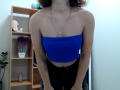 Steisii with blue tube top, showing her tits, black pants, and hands on her thighs, wearing a necklace, in a room with a wooden shelf and plants.