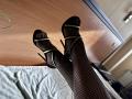Tisia's benen, bedekt met zwarte visnetkousen en hoge hakken, met haar tenen omhoog gericht, waardoor haar open sandalen en groene bandjes zichtbaar zijn. Haar voeten zijn lichtjes gekruist..
