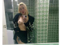 CharlotieRosse, con cabello rubio, sin sostén, mostrando pequeños senos, vestida con una chaqueta y falda negras, sosteniendo un teléfono en un baño con azulejos verdes, con un tatuaje visible en su muslo derecho..