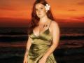 Rubbi with red hair, green satin dress, white flower in hair, tattoos on chest and arm, standing on beach at sunset, showing her tits.