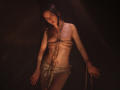 TriciaBarnes with rope bound to her chest, covering her small tits, and a cloth over her pussy, standing in dim light, looking seductively at the camera.