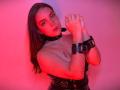 Katarina Novak met een rood-roze huidtint, handboeien, een zwarte leren choker en banden, een tatoeage op de borst, lang bruin haar, een serieuze uitdrukking, die naar de camera kijkt, met ontbloot bovenlijf..
