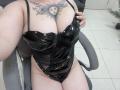 La combinaison intégrale en latex noir brillant de DiLaria, avec des découpes sur les côtés qui laissent entrevoir ses seins et ses parties intimes, est assise sur une chaise grise. Elle tient sa combinaison, montrant son tatouage de soleil..