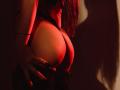 JaneWow's huge tits are highlighted by red lighting, her hands with long black nails gripping them from behind, her long red hair falling over her shoulder.