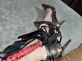 GeraNia's leg with shiny bronze high heels, black straps, and a red leather flog with black leather handles, lying on a gray suede surface.