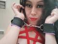 KendraSimons with red bondage straps across her tits, wearing black cuffs with purple edges, and a metal chain between them, has red lipstick and wavy dark hair.