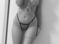 SugarrX with a flat stomach, tied ribbed shirt, black thong with floral pattern, belly button piercing, tattoos on thighs, and arms slightly raised. Black and white image.