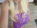 ArianaBlonde with long blonde hair wearing a purple, see-through dress, showing her tits and tattoos on her chest and arm, standing on wooden floor.