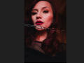 AyshaSmith with red lipstick, nose ring, and chain in mouth, dark wavy hair, black sheer top, serious expression, centered in frame.