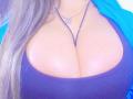 Les grandes seins brillants et bronzés de GilliSkquirtty, vêtue d'un haut noir, avec un collier violet et de longs cheveux blonds, portant une veste bleue..