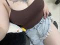 DiLaria with big, round tits in a brown, spaghetti-strap top, and frayed, light-blue, sparkly shorts, sitting on a black and white plush toy. Her red nails are visible.