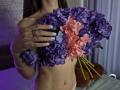 TenerliRize holding a bouquet of purple and pink flowers, covering her tits with her hand, white nails, silver ring, belly button piercing, in a dimly lit room.