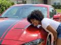 RoldanStefy leans on a red Mustang with black stripes, smiling with tongue out, wearing a white tee and denim shorts, wet car reflecting light.