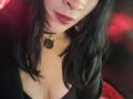IssisDiosa with black hair, medium tits, and red lipstick, wearing a black choker with a cat pendant, in a low-cut top, smiling slightly.