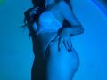 FlyFreedom with long brown hair, white lace bra, and matching panties, blue-lit, showing her ass, hand on her hip, red nails, standing in profile, touching her bra.