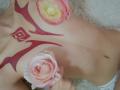 AmeliaMaiv's tits with red tribal tattoos, holding pink roses on her chest and belly, wearing white lace panties, lying on a fluffy surface.