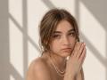 BessBetak with messy brown hair, green eyes, and a pearl necklace, hands gently touching her face, looking slightly sad or thoughtful, topless with subtle shadows on her skin.