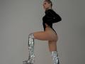 MilagrosEdmisten, a fit woman with blonde hair, stands in profile, one leg raised, wearing a black top and shiny silver knee-high boots. She's naked below the waist, showing her ass.