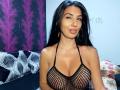 BrunetteBabe69 with long black hair, wearing a black fishnet top, showing her large tits, pouty lips, and gold earrings, in a bedroom with purple and black patterns.
