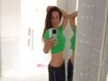 ViktoriiaLu with long brown hair, green crop top, and black pants, standing in front of mirror, arms raised, holding phone, showing her flat stomach and small tits.