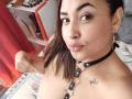 MirandaDreams with brown hair, wearing a black choker with metal rings, and a tattoo of a small butterfly on her right shoulder, showing her tits in a selfie.